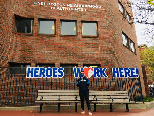 A person stands in front of the East Boston Neighborhood Health Center wearing a mask in front of a sign that says Heroes Work Here,