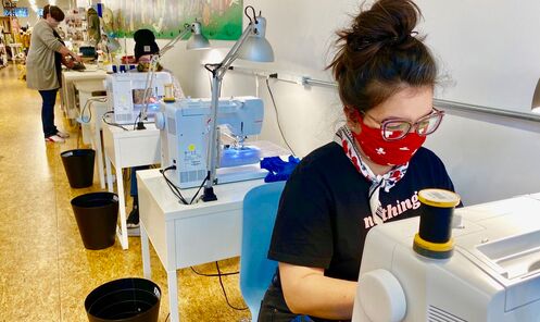 A women wearing a red bandana as a face mask sews at the head of a row of sewing machines.