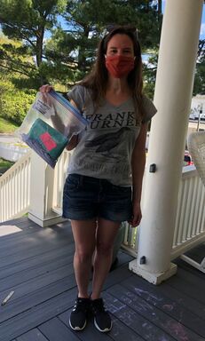 A volunteer wearing shorts and a t-shirt holds a bag of masks on a porch.