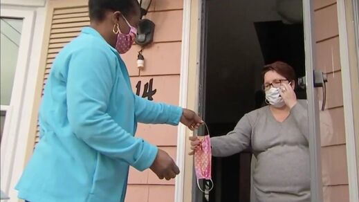 A woman hands a fabric face mask to another woman at her home. 